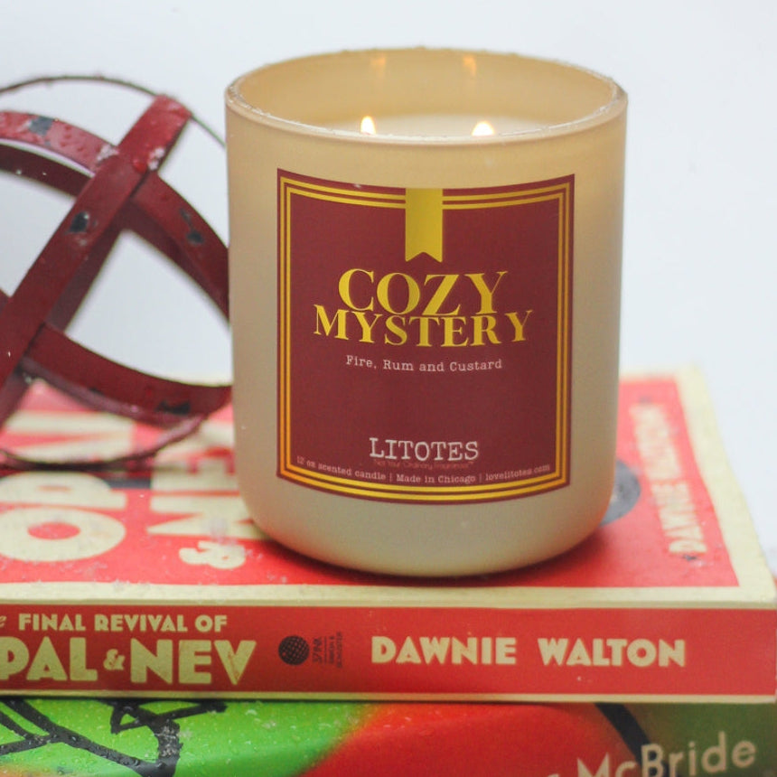 Candle labeled 'Cozy Mystery' on top of books with a red decorative object in the background.