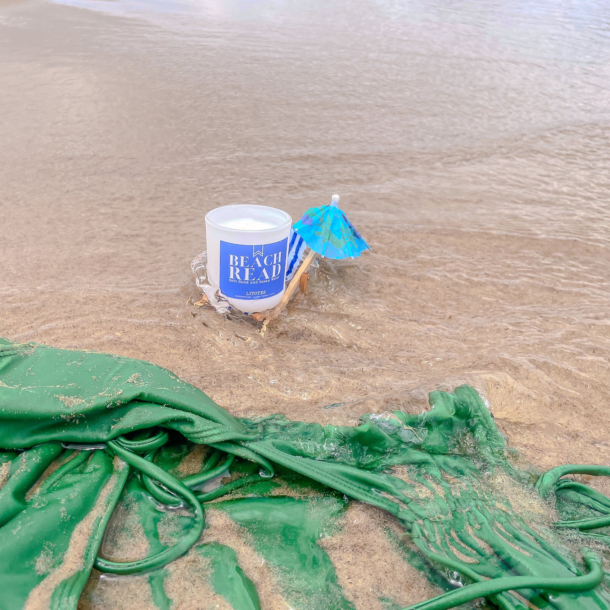 Candle labeled 'Beach Read' with a small umbrella and green swimsuit on sand.
