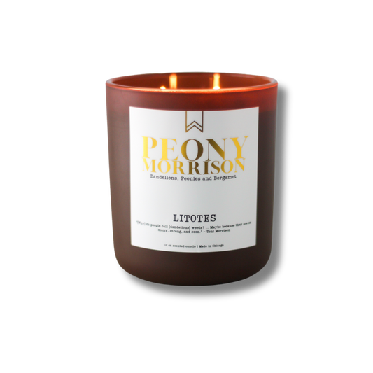 A soy wax candle with a label that reads 'Peony Morrison', with a lit flame inside a dark glass jar.